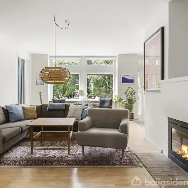 En stue med en grå hjørnesofa og lænestol, placeret på et tæppe. Der er et sofabord i midten. En pejs til højre brænder. Store vinduer viser en have.