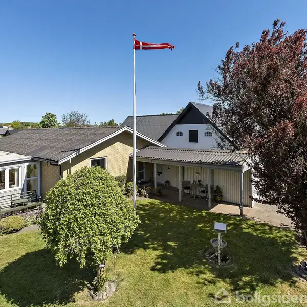 Et gult hus med en carport og en dansk flagstang i haven. Træer og buske omkranser huset i et solrigt miljø.
