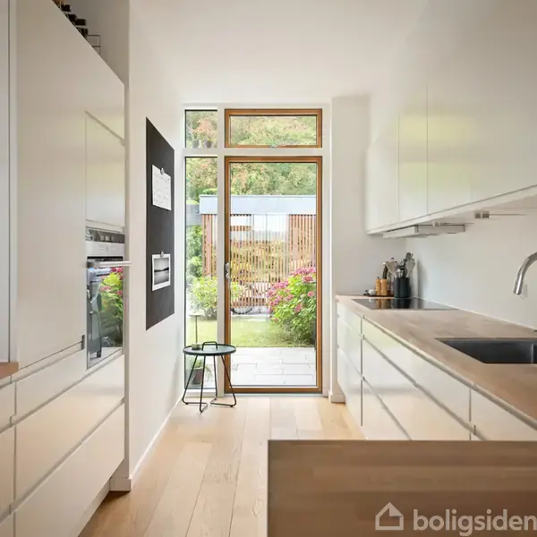 Et moderne, hvidt køkken med træbordplader. En vask og kogeplade ses. Store vinduer for enden åbner op til en have med blomster og buske. En sort skammel står ved vinduet.
