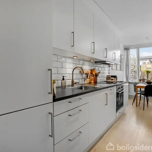 Et hvidt køkken med sorte detaljer viser en vask og komfur. I baggrunden står et bord ved et vindue, der giver udsigt til træer udenfor.