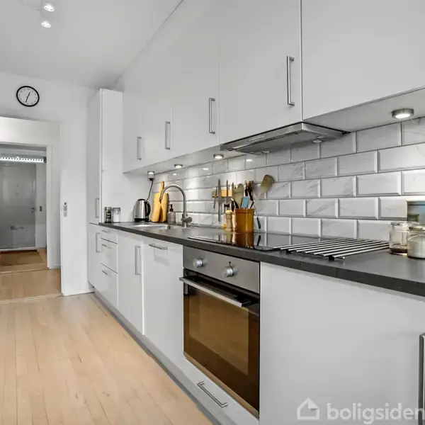 Et moderne køkken med hvide skabe og stålgreb, en indbygget ovn og kogeplade. Væggen har hvide fliser. Til venstre hænger forklæder. En dør åbner til en gang.