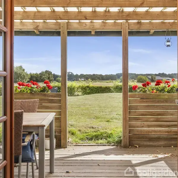 Dobbelt glasdør åbner ud til en terrasse med træoverdækning. Et bord med stole står ved siden af blomsterkasser med røde blomster. Udsigt til en grøn mark under blå himmel.