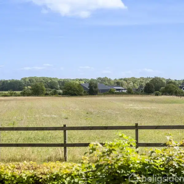 En græsmark strækker sig med et træhegn i forgrunden. I baggrunden ses huse og skov under en klar blå himmel med få skyer.
