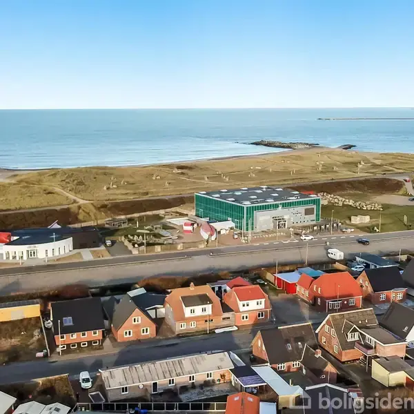 Bygning med glasfacade, står ved kystlinje omgivet af græs, hav og klitter i baggrunden, mens farverige huse er placeret i forgrunden ved vejen.