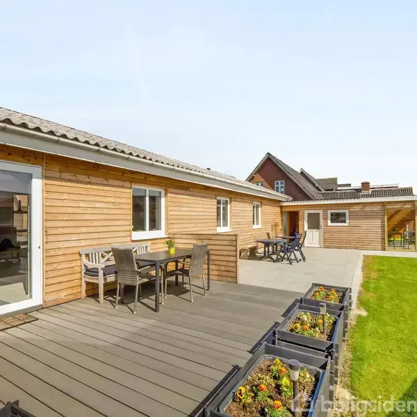 Træhus med terrasse i forgrunden og græsplæne i baggrunden. Terrassen har spisebord og stole. Blomsterkasser med planter langs kanten. Klar himmel.