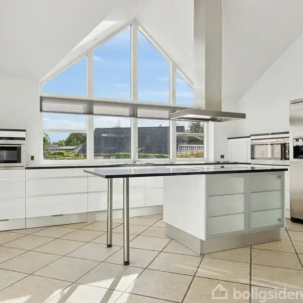 Et lyst køkken med en stor ø i midten, omkranset af hvide skabe og apparater. Store vinduer lader naturligt lys strømme ind, med udsigt til grønne omgivelser udenfor.