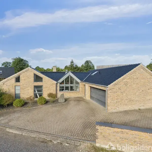 Et moderne murstenshus med blåligt tag står på en flisebelagt indkørsel omgivet af grønne buske og træer, beliggende i et boligkvarter med klart blå himmel i baggrunden.