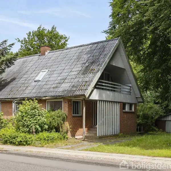 Et rødt murstenshus med skråt tag står på en stille vej omgivet af frodige træer og buske, med en græsplæne foran og en lille garage ved siden af.
