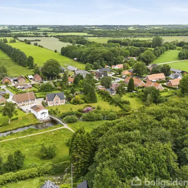 Landsbyhuse med røde tage ligger spredt mellem grønne marker og skove. Et vandløb og en bro er synlige i nærheden, mens det omgivende landskab strækker sig på en klar dag.