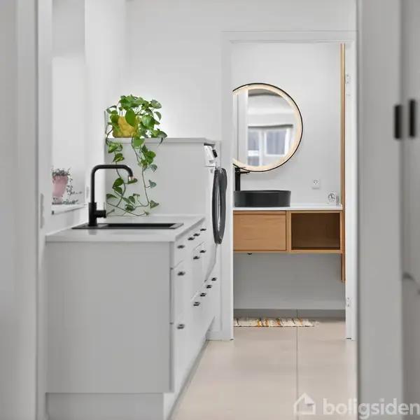 Køkken med hvide skabe og sort håndvask, prydet med en grøn plante, fører til et badeværelse med rundt spejl og vask, i et lyst og rent miljø.