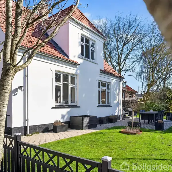 Hvidt hus med rødt tegltag står stille i en velholdt have. Der er en terrasse med havemøbler og grøn græsplæne omgivet af træer og en sort stakit.