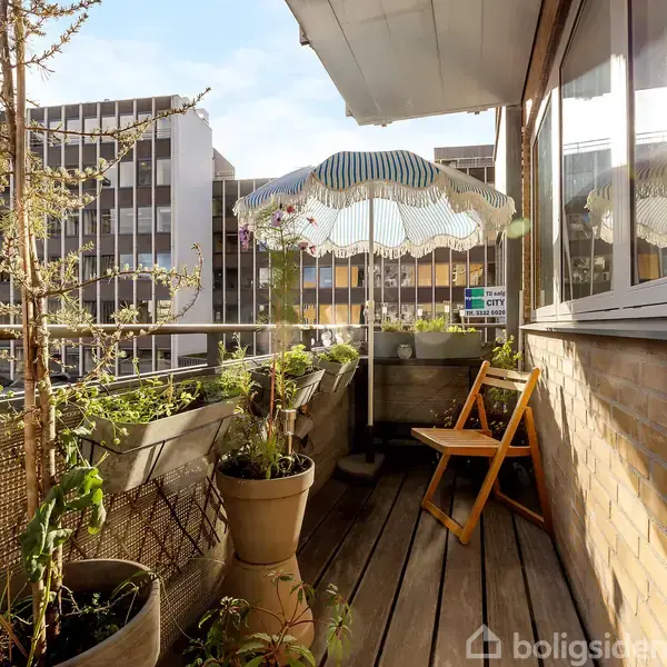 En altan med potteplanter, foldestol og parasol, placeret i en urban setting med udsigt til omkringliggende bygninger. Sollys reflekterer ind gennem glasvæggen.