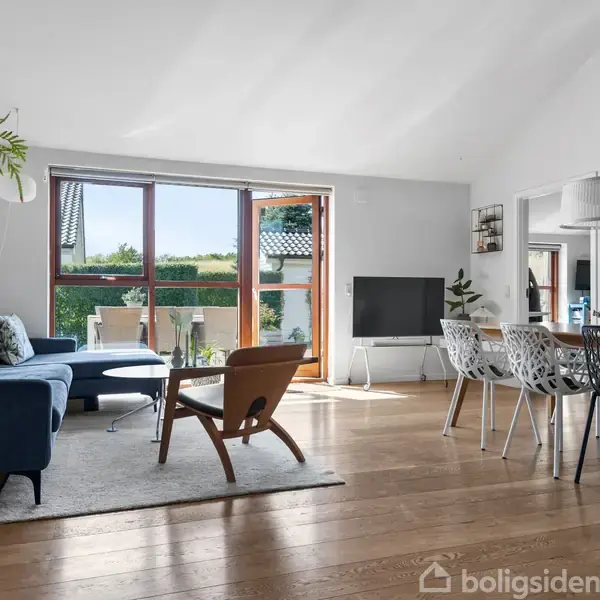 Stue med blå sofa og lænestol på gulvtæppe, stor træspisebord omgivet af hvide stole, store vinduer til terrasse og grøn udsigt, moderne interiør med planter.