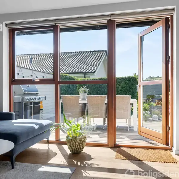 Stue med blå sofa og bord, åbne glasskydedøre fører til terrasse med grill og spisebord. Lyset strømmer ind fra haven og skaber en lys atmosfære.