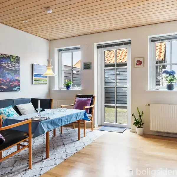 Stue med spisebord og stole, dækket med blå dug. Væggene har kunstmalerier. To vinduer og en glasdør slipper lys ind, planter på gulvet og en kommode til højre.