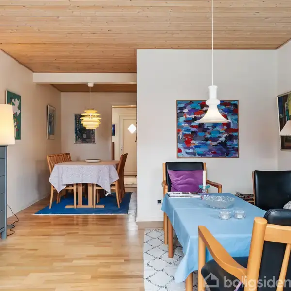 Et lyst køkken-alrum med træmøbler og grå bordplade. Køkkenet har hvide skabe, komfur og er omgivet af brune fliser. Spiseområdet har en bordlampe og lysindfald fra vinduet.