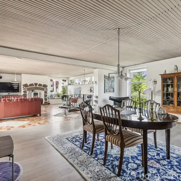 Et spisestuebord med stole står på et tæppe i et stort, lyst opholdsrum. I baggrunden ses en rød sofa og pejs, med grønne planter ved vinduerne.