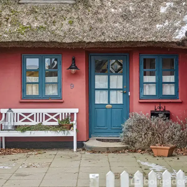 Et hyggeligt, stråtækt hus har en blå dør og vinduer, flankeret af en hvid bænk og planter. Tegnet "9" hænger ved siden af døren. Vejret ser koldt ud.