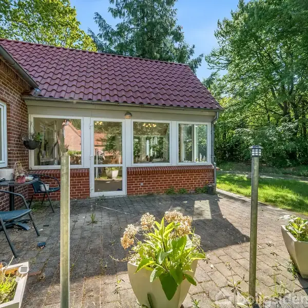Et rødt murstenhus med en veranda med store vinduer åbner ud mod en terrasse med blomsterkrukker og havemøbler. Omkring huset er der en frodig grøn have med træer.