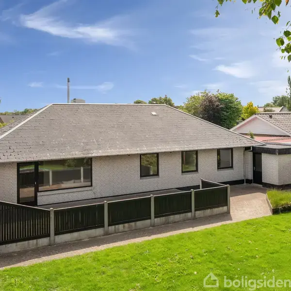 Huset, en grå enkelt-etagers bygning med skråtag, står i en grøn have. En indhegnet terrasse er foran, og baggrunden viser nabohuse og træer under en blå himmel.