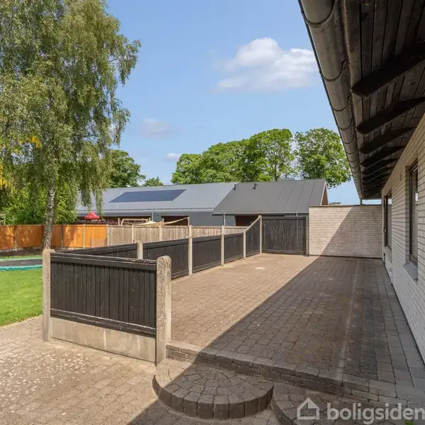 En stor terrasse foran en hvid murstensbygning viser fliser og lave hegn. Til venstre er en græsplæne med træer og i baggrunden ses en anden bygning og blå himmel.