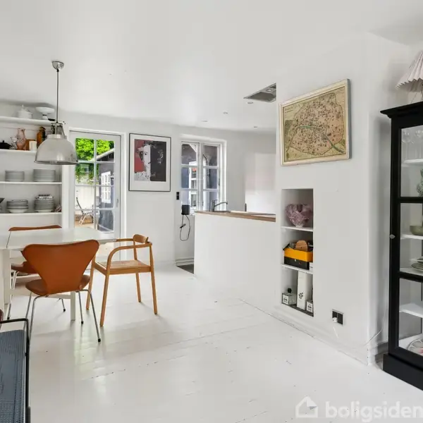 En lys stue med hvide møbler indeholder reoler, en spisebordsgruppe med træstole, en skænk med pyntegenstande og et vitrineskab fyldt med porcelæn. En åben dør fører udenfor.