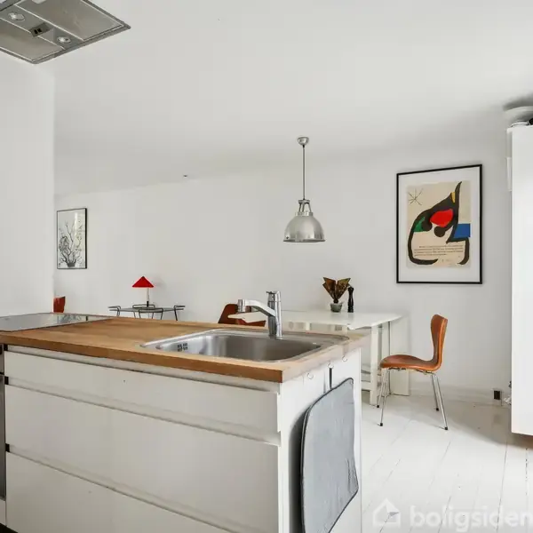 Et moderne køkken med en hvid køkkenø indeholder en vask og kogeplade. Rummet er lyst med minimal indretning; plakater på væggen, spisebord med stole og en pendellampe i baggrunden.