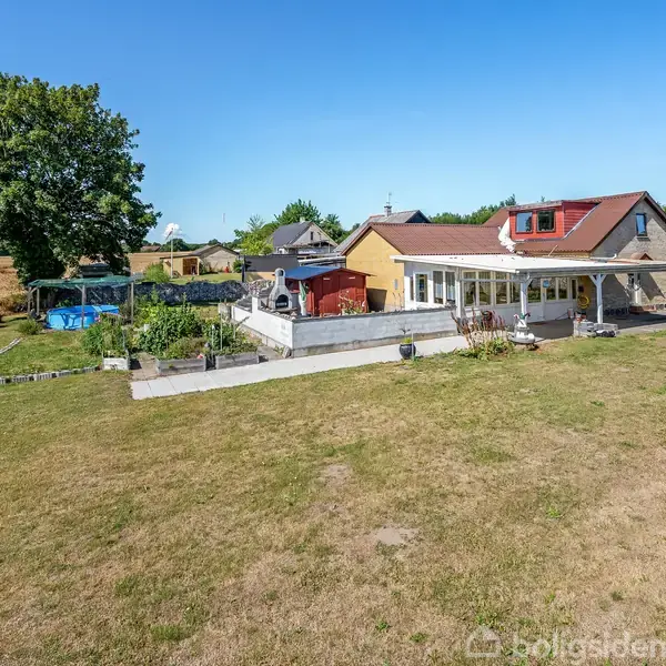 Et gult hus med stor terrasse omgivet af græsplæne og træer. En swimmingpool er placeret til venstre ved siden af en have. Klar blå himmel i baggrunden.