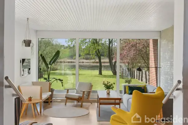 En lys stue med moderne møbler står stille. Store vinduer i baggrunden viser en have med grønne træer og græs. Bordet har en vase med blomster, mens stole og sofaer omkranser rummet.