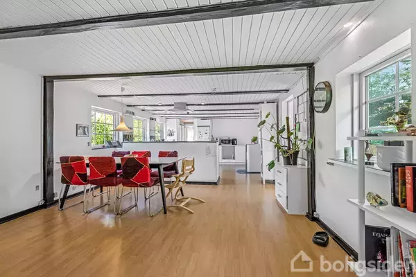 Spisebord med røde stole står i en lys spisestue. Det åbne rum har trægulve, hvide vægge og loft med synlige bjælker. Køkkenet ses i baggrunden. En plante står ved en skænk, og et vindue giver udsigt til haven udenfor.