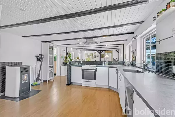 Køkken med hvide skabe og en central komfur, rene træbjælker i loftet, lyst trægulv, en moderne brændeovn til venstre og store vinduer, der giver naturligt lys.