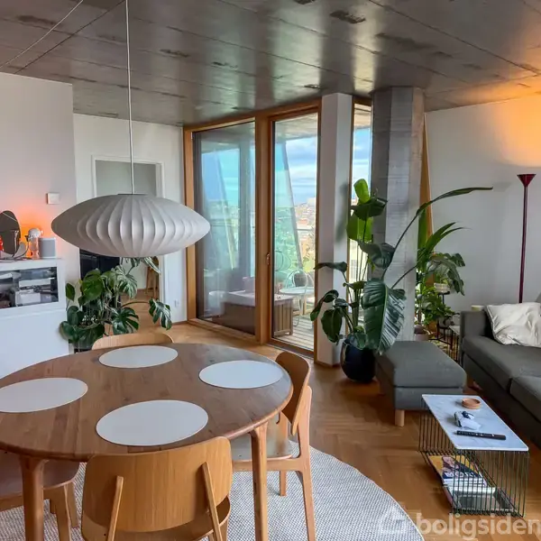 Et rundt træbord står i en moderne stue med stole omkring det. Store planter pynter rummet, der er desuden en sofa, glasdøre til altan og en stor loftslampe.