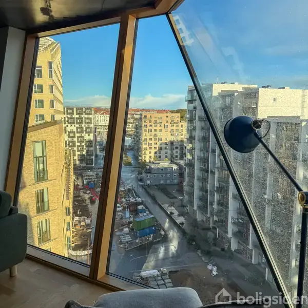 Udsigt fra en lejlighedsvindue viser høje, moderne bygninger i et byområde. En bordlampe står ved vinduet, mens nederst ses en gård med byggepladsaktiviteter.