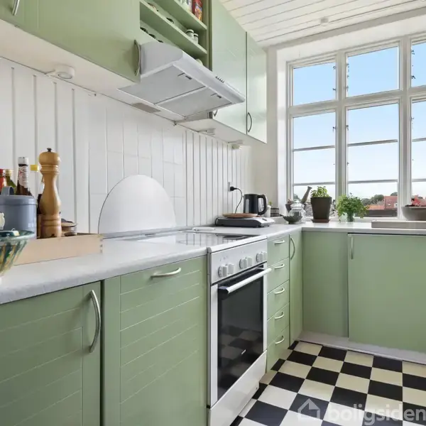 Grønt køkken med ovn og kogeplade på en hvid bordplade. Sort-hvidternet gulv. Flasker og urter er placeret på bordet. Store vinduer giver udsigt og naturligt lys.