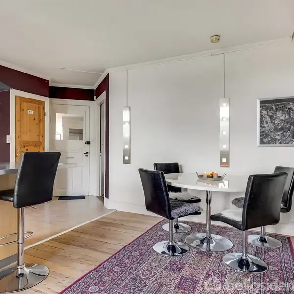 En spiseplads med et rundt bord og fire sorte læderstole på en rødligt tæppe. Baggrunden har hvide vægge, et sort-hvidt billede, og en lille, moderne barområde til venstre.