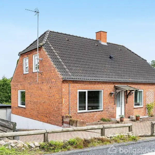 Et rødt murstenshus har en antenne på taget og en hvid hoveddør med et lille halvtag. Det ligger ved en vej omgivet af træer og buske.