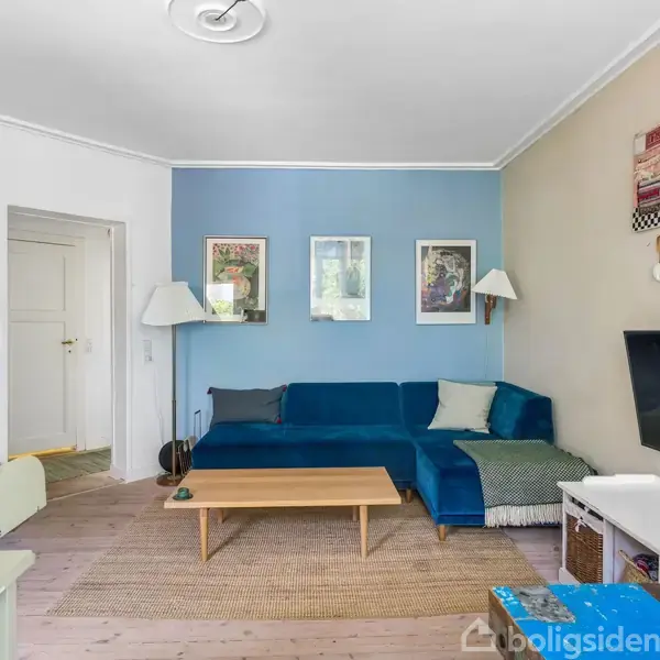 En stue med en blå sofa står i hjørnet op ad en blå væg, dekoreret med tre billeder. Foran sofabord står en tv-skærm. Blomster står på bordet til højre.