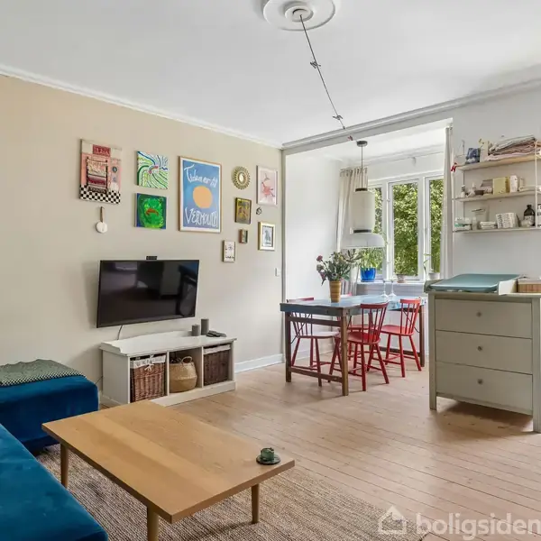 Stue med blå sofa, vægophængt fjernsyn og trækaffebord. Spisebord med røde stole ved vindue. Hylder og en kommode ses ved siden af en åben altandør. Vægpynt inkluderer flere kunstbilleder.