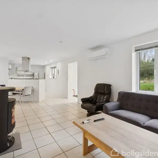 Stue med mørk sofa og lænestol ved træbord. Brændeovn til venstre. Flisegulv leder til køkken med komfur og emhætte. Vindue viser grøn udsigt.