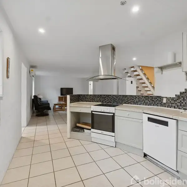 Køkken med hvide skabe har en opvaskemaskine og ovn. Køkkenet åbner ind til en stue med trappe og flisegulv. Et vindue til venstre, stue til højre. Ingen tekst.