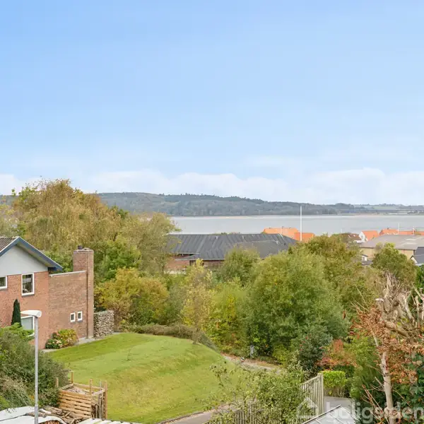 Et hus med rødt murværk står i en have med græs og buske, omgivet af andre huse og træer, med udsigt over en fjord eller sø i baggrunden.