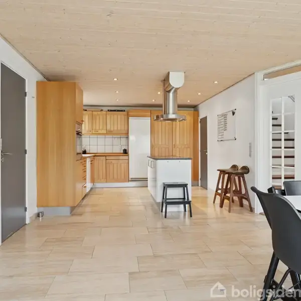 Et rummeligt køkken med træskabe og en ø med en indbygget emhætte. Køkkenet åbner ind til en spisestue med sorte stole. Lyse gulvfliser og en trappe i baggrunden.