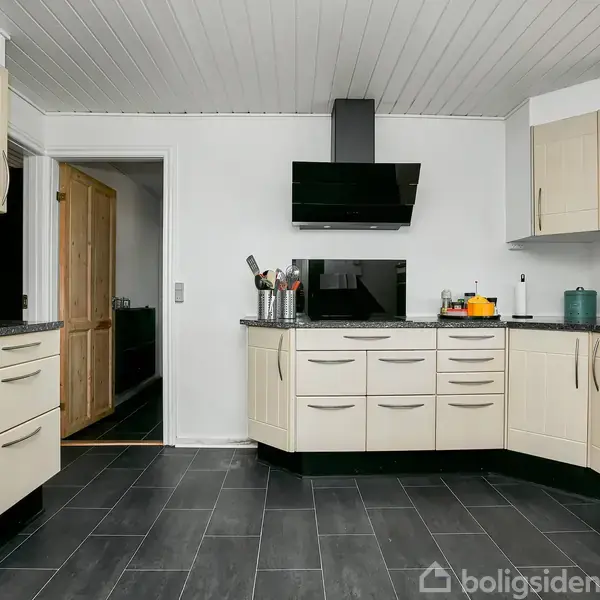 Køkken med lyse skabe og sort bordplade; apparater og knivholder står på bordet. Sort flisegulv og hvide vægge. Åben dør fører til et tilstødende rum.