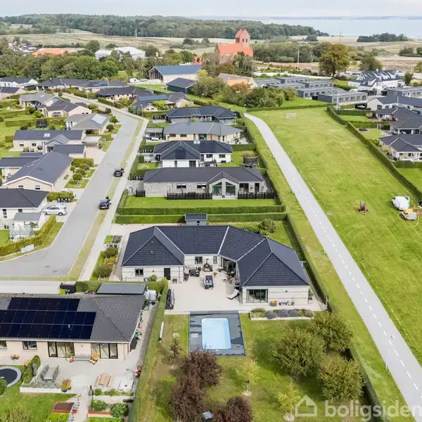 En række moderne parcelhuse med sorte tage ligger langs stille veje. Nogle haver har pools. En vej med græsarealer til højre og en kirke er synlige i baggrunden.