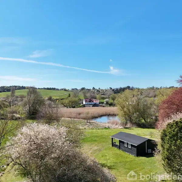 En sort træhytte står stille på en grøn eng omgivet af blomstrende træer og en lille sø, der reflekterer en klar blå himmel. En hvid villa ses i baggrunden.