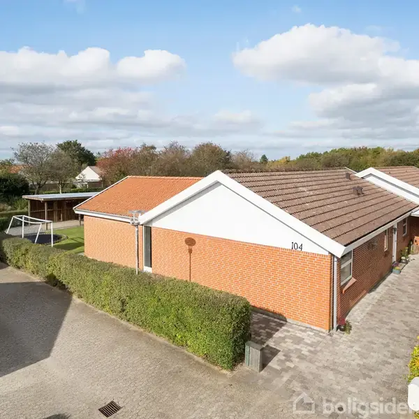 Et rødt murstenshus med hvide gavle står stille omkranset af hække og en flisebelagt parkeringsplads. Nummeret "104" er synligt på gavlen. I baggrunden ses et træskur.