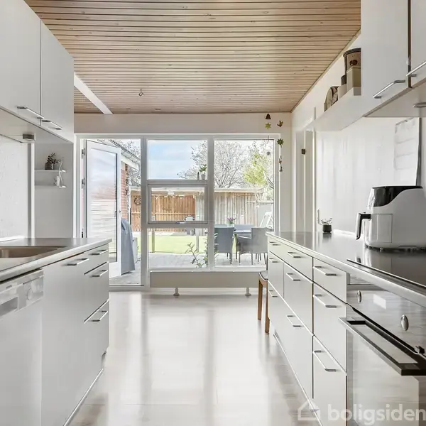 Køkken med hvide skabe og stålgreb, indeholder indbygget ovn, vask og knivrække på væg. Stor glasdør åbner ud mod en terrasse og have i baggrunden.