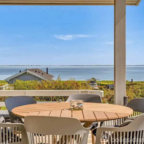 Rundt bord med seks stole står på en overdækket terrasse med udsigt over havet og kystnære huse. Solrigt vejr og blå himmel i baggrunden.