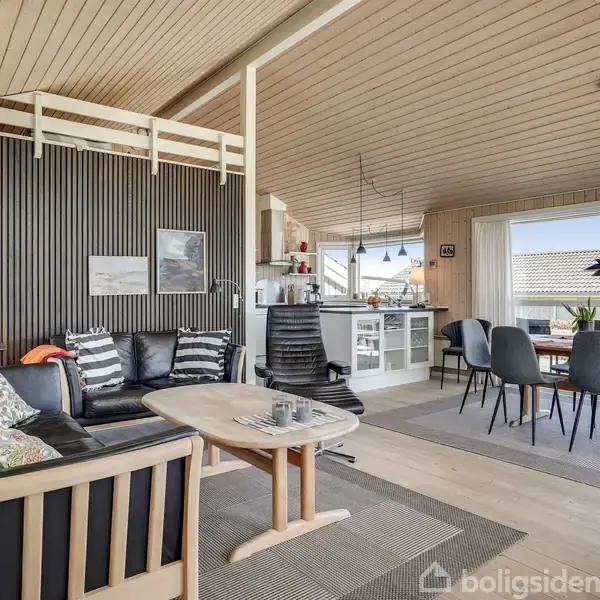 Stue med sort lædersofa og sofabord, i forbindelse med et åbent køkken med spisebord. Højt til loftet med træpaneler, store vinduer med udsigt til tage, indrettet med moderne møbler.