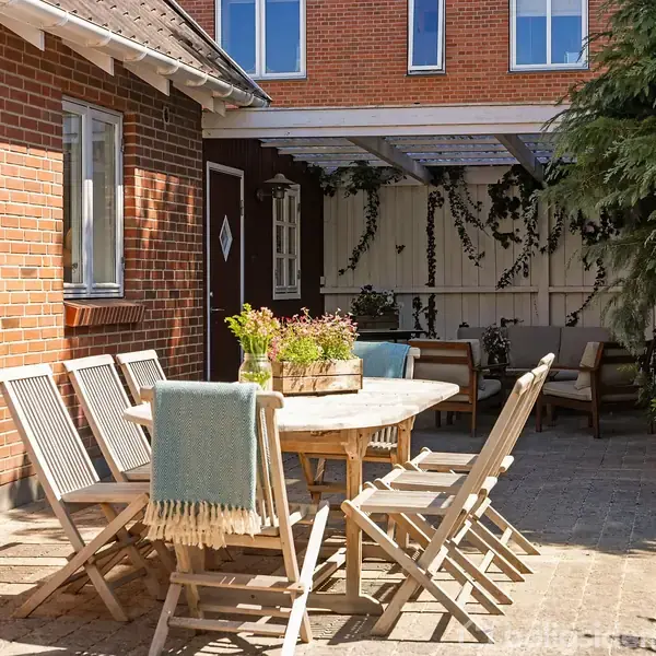 Et bord med træstole står på en flisebelagt terrasse ved siden af et murstenshus. Et tæppe hænger på en stol, og planter dekorerer området med overdækket siddeplads i baggrunden.